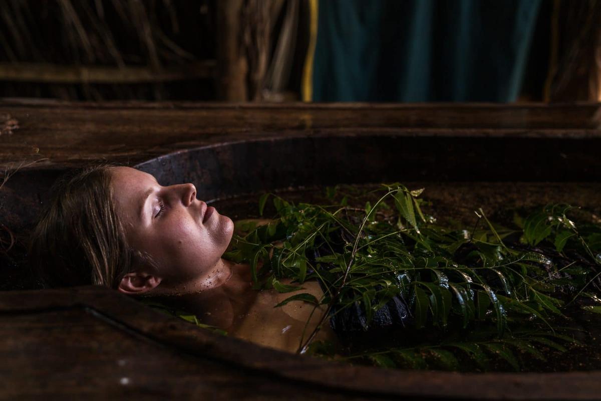 A massage therapist works on a guest lying on a wooden table in an open-air thatched pavilion draped with purple and tie-dyed fabrics, jungle boulders behind.