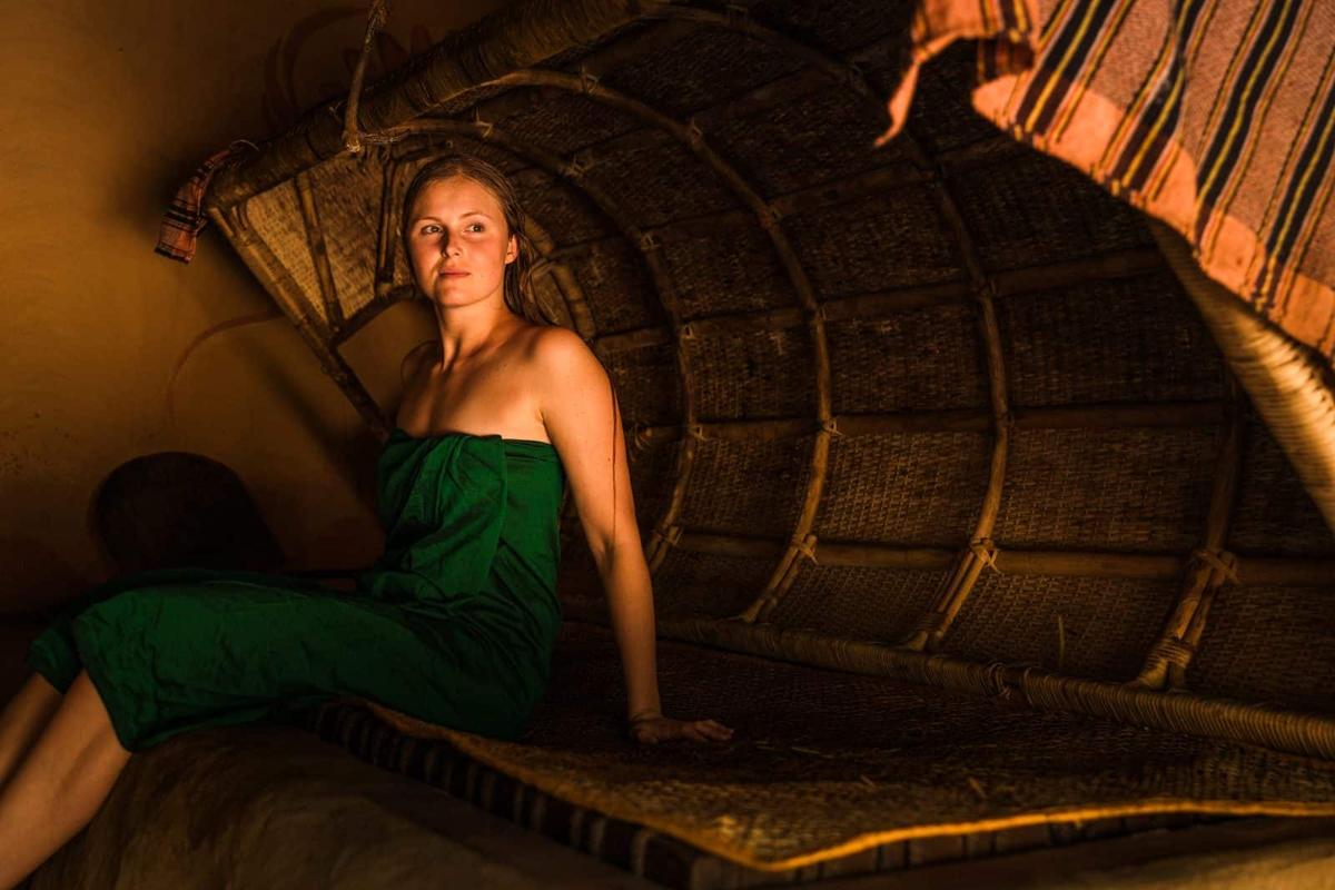 A guest wrapped in green cloth sits on the woven mat of the steam bath, its rattan hood raised overhead