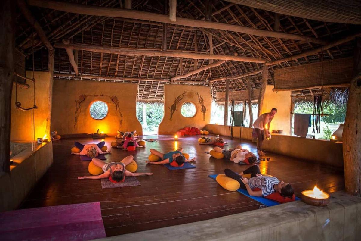 A candlelit restorative yoga class at dusk inside the open-air thatched shala, guests lying over bolsters on mats while a teacher adjusts one student.