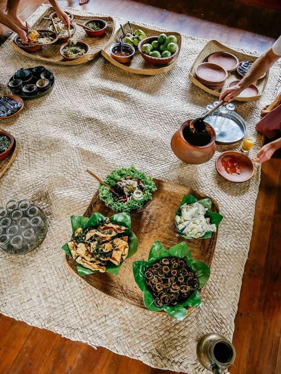 Dinner laid out on banana leaves in woven rattan trays across pale rush mats, hands reaching in to serve from clay pots