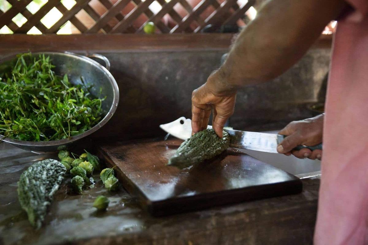 Weathered hands slicing a bitter gourd on a dark wooden board in the open kitchen, a colander of just-picked greens beside