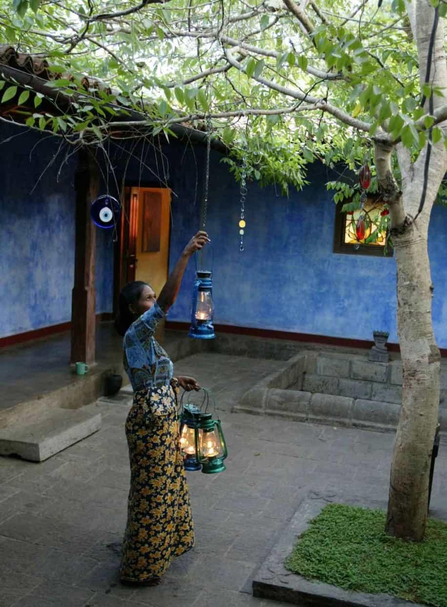 A woman in a batik sarong hanging a lit blue-glass oil lantern from a tree in the indigo-walled courtyard at dusk