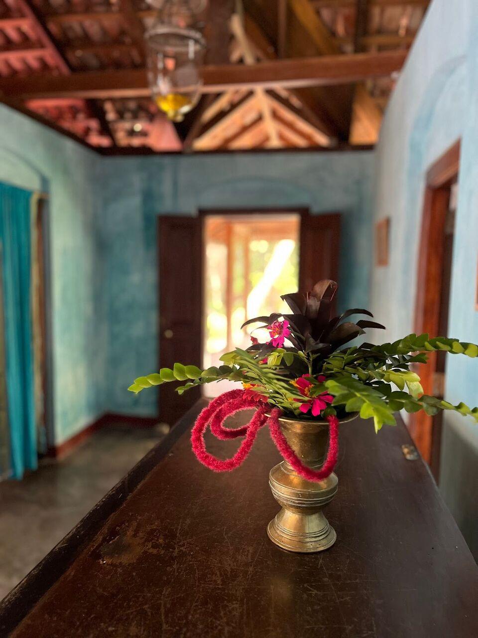 A brass vase of red flowers on a dark wooden table in a turquoise room, a sunlit doorway beyond