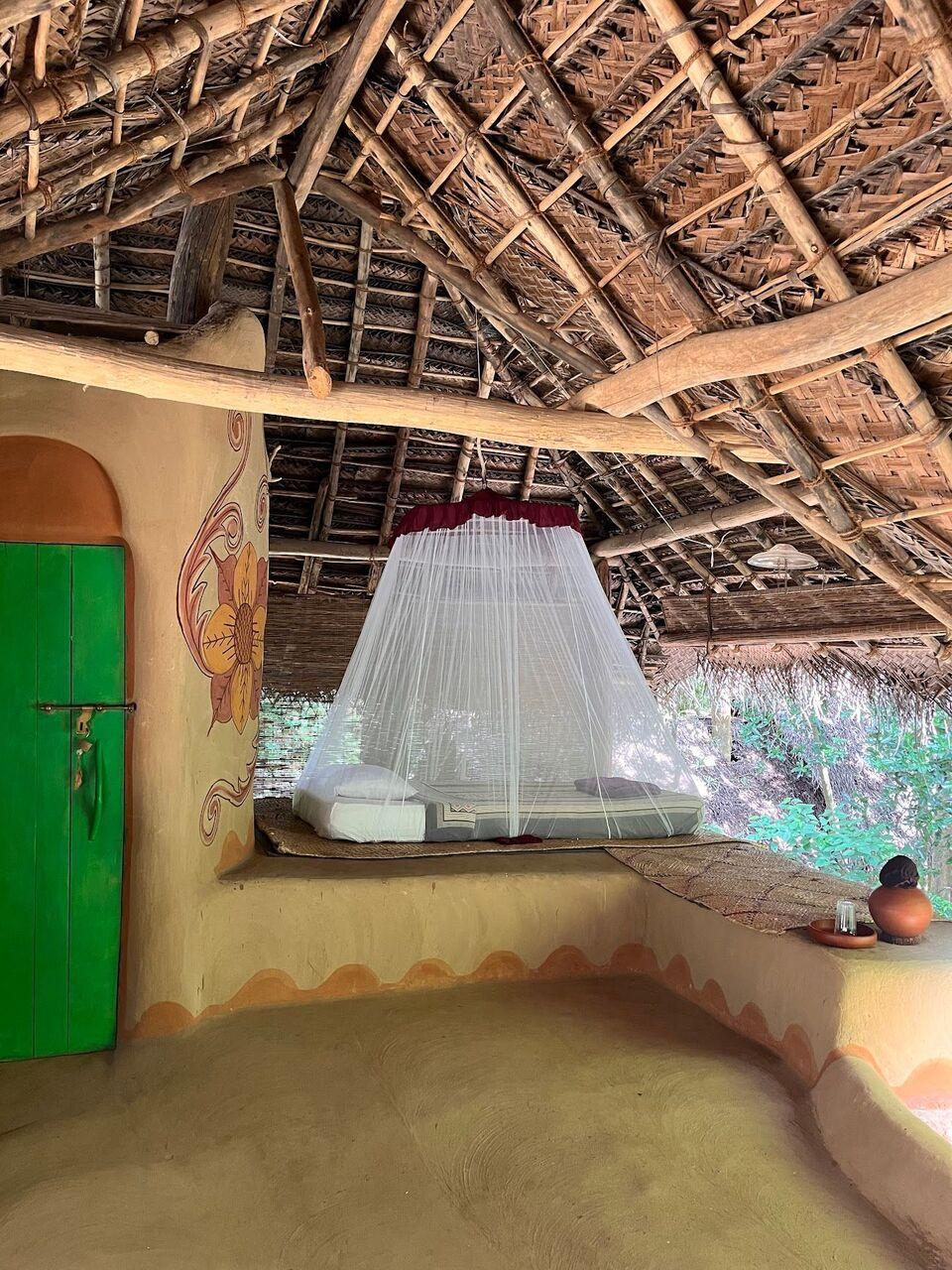 Inside a mud hut: a sculpted bed under a white mosquito net, a green door and a woven palm-leaf roof