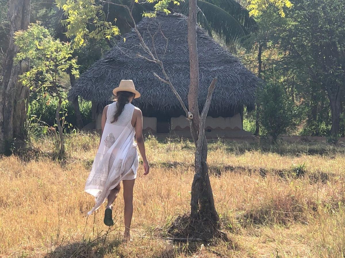 A woman in a white dress and straw hat walking through dry grass towards a thatched hut