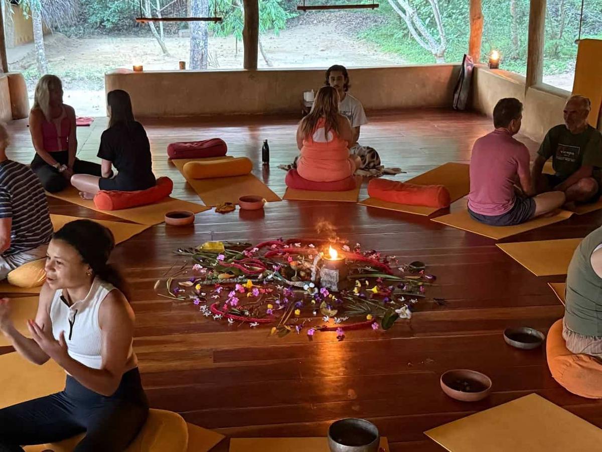 A shaven-headed yoga teacher in white sits cross-legged on a mat, gesturing mid-instruction beneath a huge banyan tree with a stone Ganesh statue, flowers and lit oil lamps behind him.