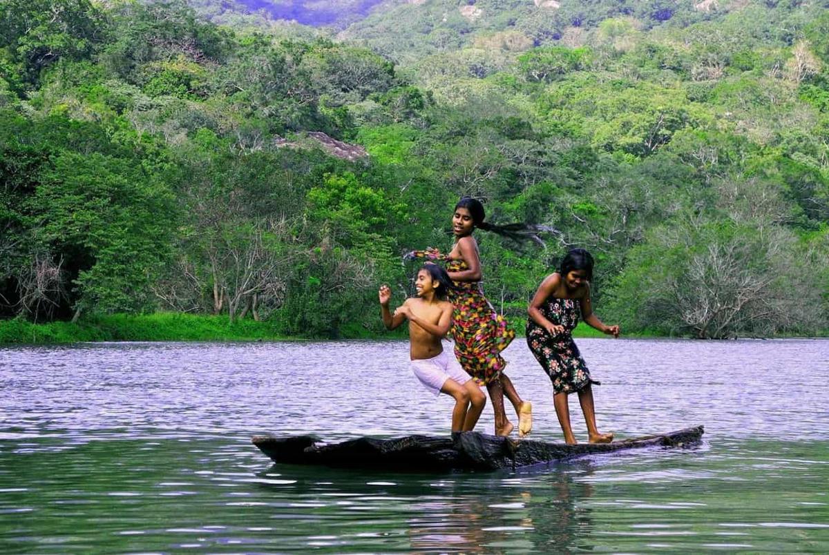 Three village children laughing and balancing on a wooden dugout canoe on the lake, jungle hillside behind
