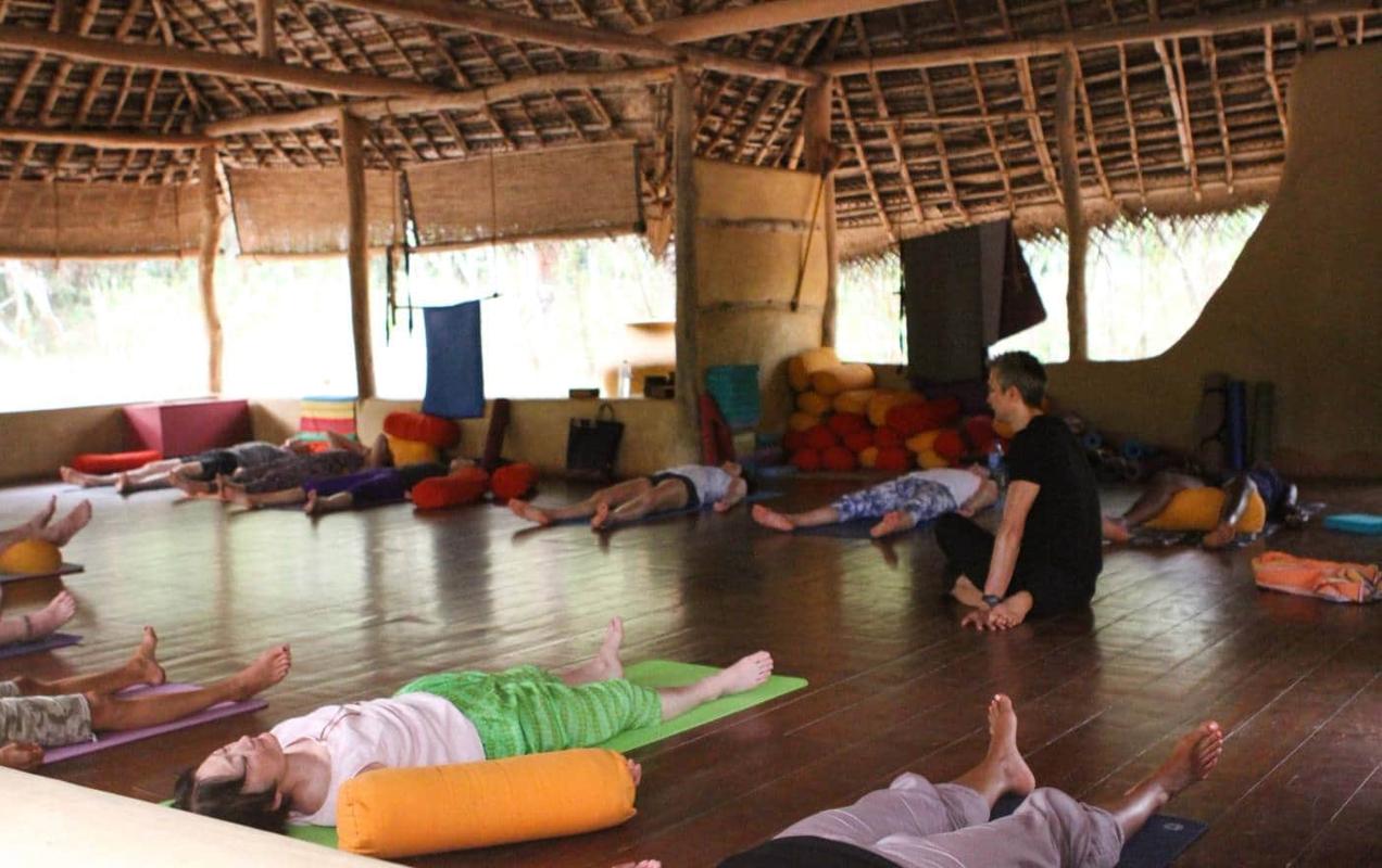 Guests resting on mats and bolsters in the open-sided shala, one sitting up among them