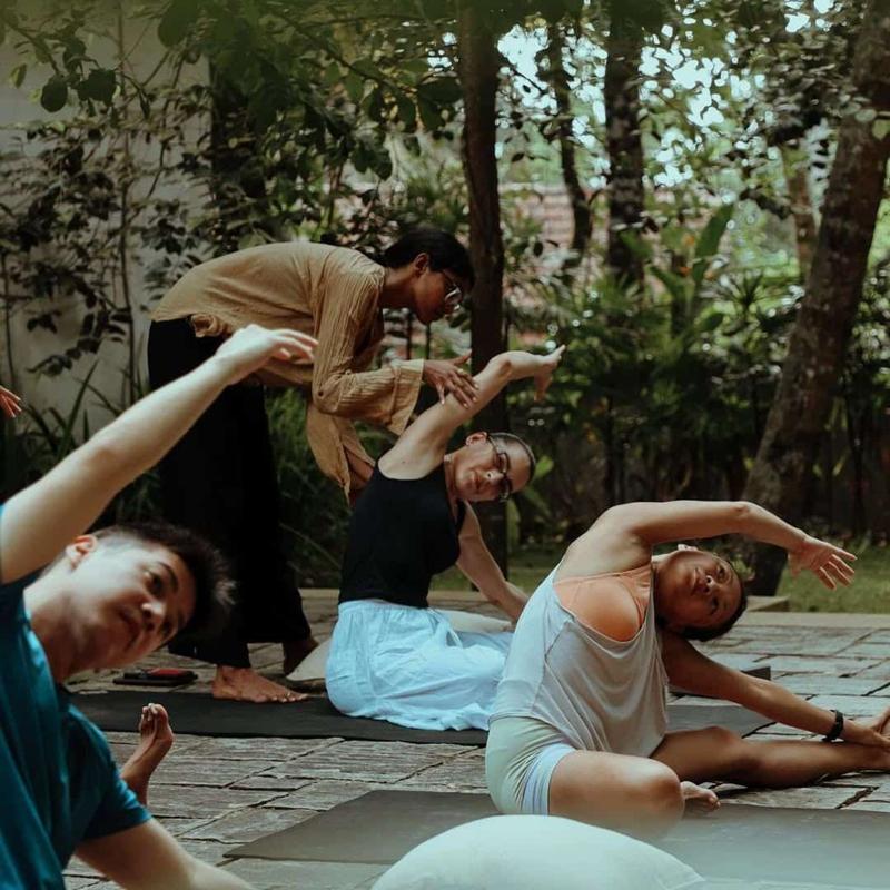 Guests in a seated side stretch on a stone terrace under the trees, one being helped into the pose