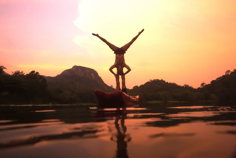 Two people performing an acro-yoga handstand balance on a boat on a still lake at sunset, with hills silhouetted behind.