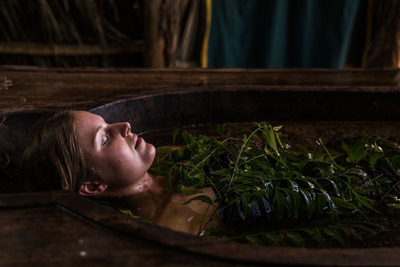 A guest resting with closed eyes in a carved wooden herbal bath, fresh neem branches floating on the dark herb-infused water, under a thatched roof