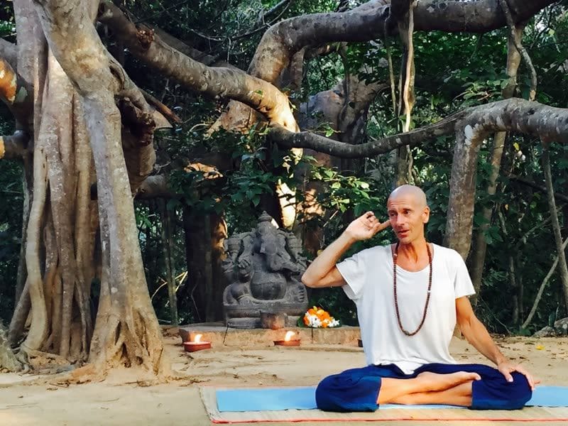 Yoga teacher seated cross-legged on a mat under a banyan tree, speaking to a class, with a stone Ganesh statue and lit oil lamps behind him.