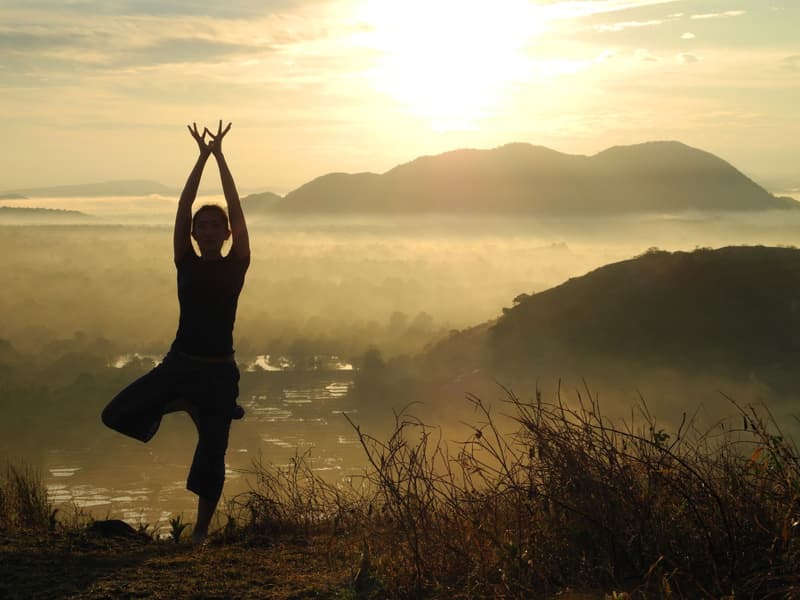 Silhouette of a person in tree pose on a hilltop at sunrise, a blanket of golden mist filling the valley over the lake and paddy below