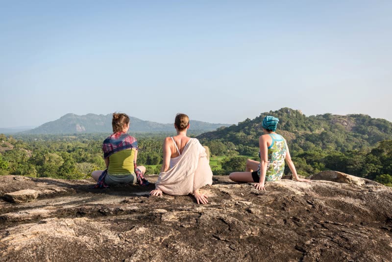 Three women sitting on a granite rock, viewed from behind, gazing over jungle and distant hills in soft golden light.