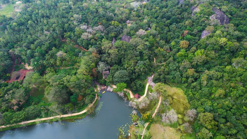 Aerial view along the lake edge: dense jungle canopy, giant granite boulders, thatched roofs hidden in the trees, a sandy path tracing the shore and canoes at a small landing