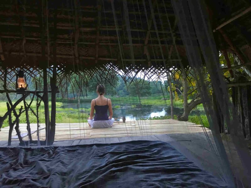 Seen from inside an open thatched hut at dim early light, a woman sits cross-legged in meditation on a bamboo deck facing the still lake, a lit hurricane lantern hanging beside her