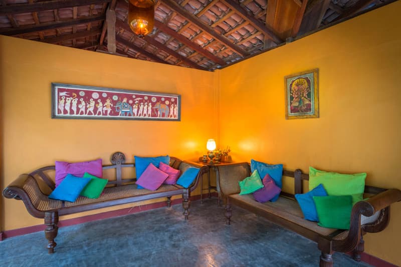 A marigold-yellow lounge under exposed clay-tile roof beams: antique cane settees piled with jewel-tone cushions, a folk-art frieze and a brass lamp aglow