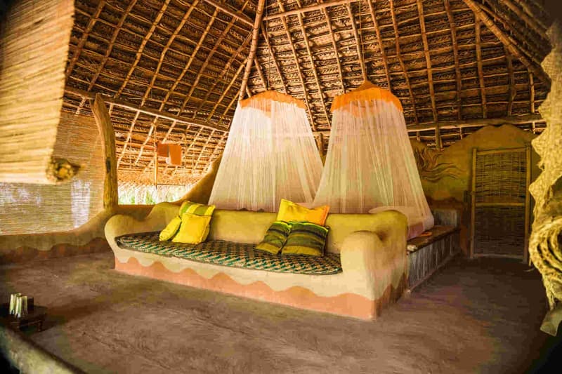 Inside a mud hut: a sculpted mud platform bed with a patterned mattress and cushions, two coned white mosquito nets hanging from the woven-palm thatch ceiling