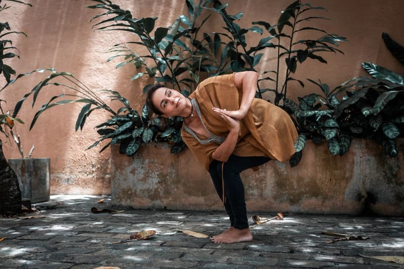 A woman in a mustard cover-up in a standing side-bend on mossy stone paving before a salmon-pink wall lined with dark tropical plants