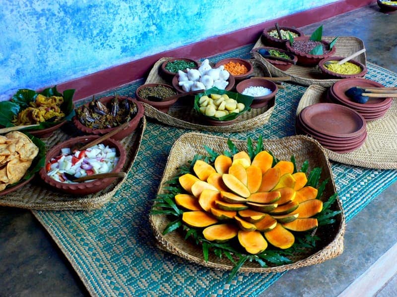 A buffet of clay bowls and woven trays against a vivid blue lime-washed wall: a spiral of cut mango and papaya at the centre, surrounded by curries, sambols and stacked terracotta plates