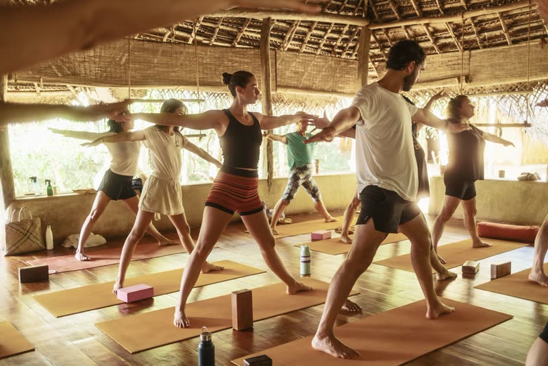 Guests holding Warrior II pose together on orange mats in Ulpotha's open-air thatched yoga shala.