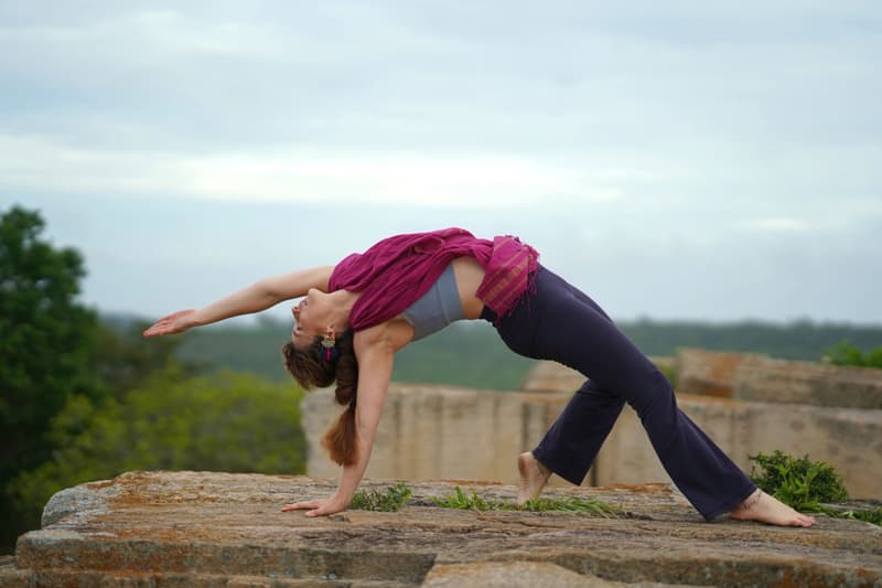 A woman in a magenta scarf and plum trousers arching into wild thing pose on a lichen-covered granite outcrop, jungle canopy below a soft overcast sky