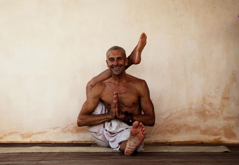 A tanned, shaven-headed yogi seated against a weathered lime-plaster wall, one leg behind his head, hands in namaste, beaming